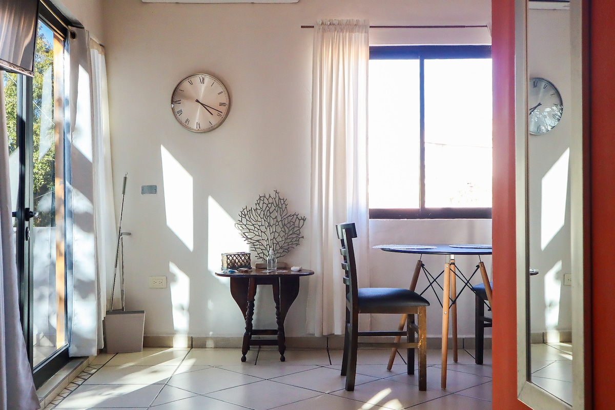 A dining area is shown featuring a round table with two chairs, complemented by a decorative tree sculpture. Large windows allow natural light to fill the space, highlighting the crisp white curtains. A wall clock is positioned above the table, adding functionality and style.