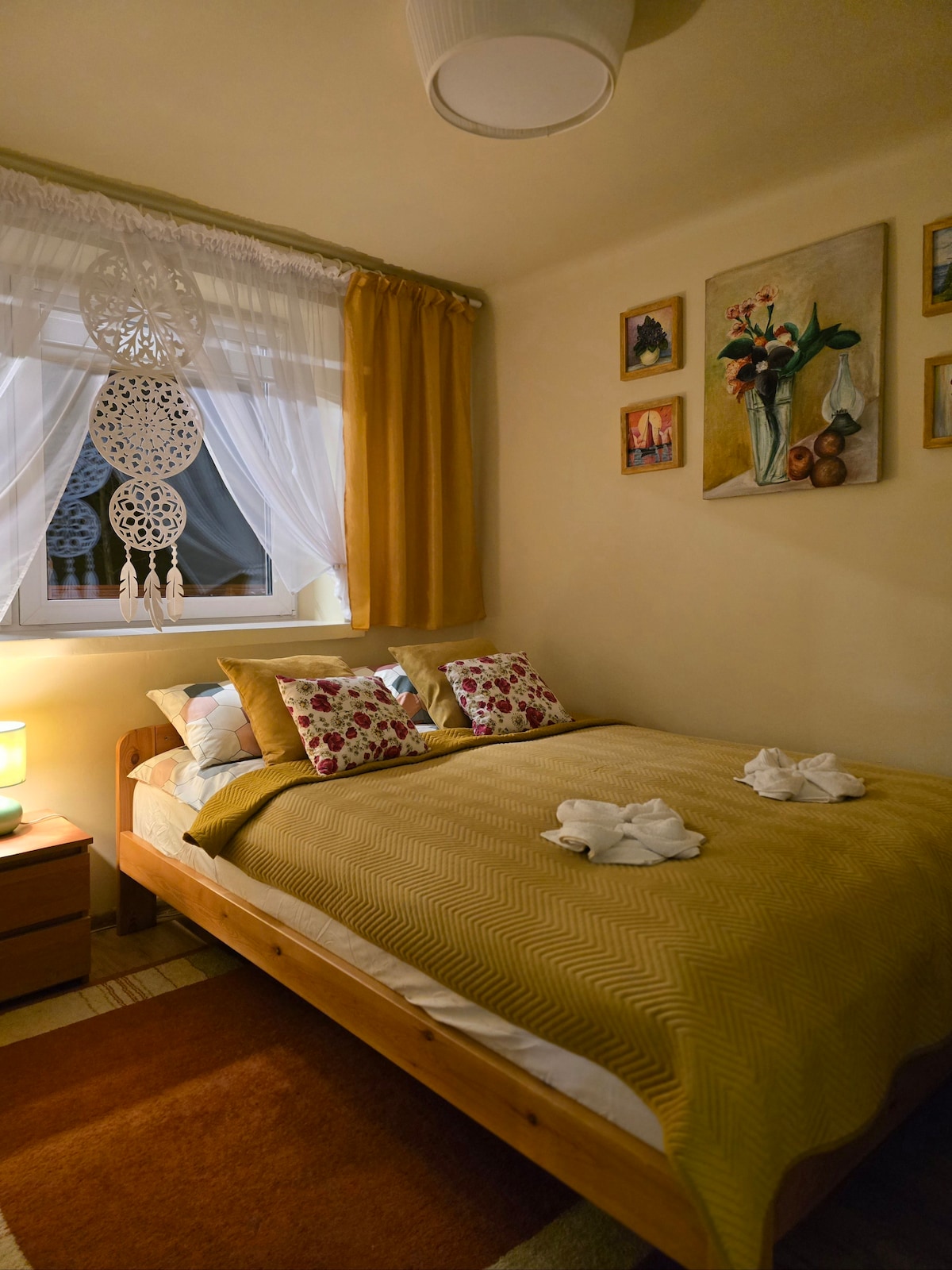 A cozy sleeping area features a spacious bed adorned with patterned bedding and decorative pillows. Natural light enters through a window dressed with white curtains. A bedside table holds a soft lamp, and artwork is displayed on the walls, enhancing the room's inviting look.