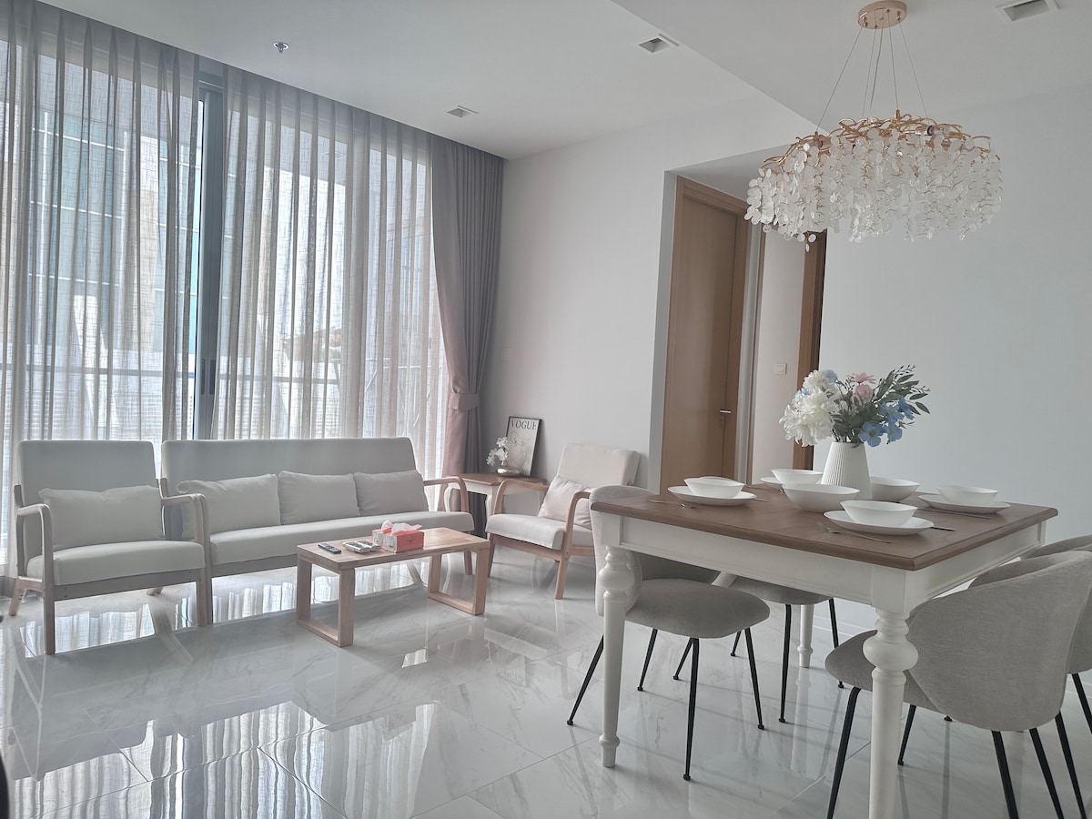 A modern living and dining area showcases a spacious layout with large windows covered by sheer curtains, allowing natural light to filter in. A stylish chandelier hangs above a wooden dining table, while comfortable seating arrangements invite relaxation.