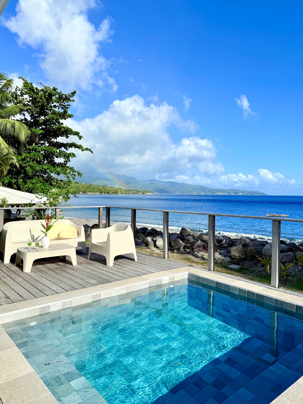 A private pool is surrounded by a wooden deck, with two light-colored lounge chairs positioned for relaxation. Lush greenery and the ocean are visible in the background, under a clear blue sky dotted with clouds. The tranquil setting invites calm and leisure.