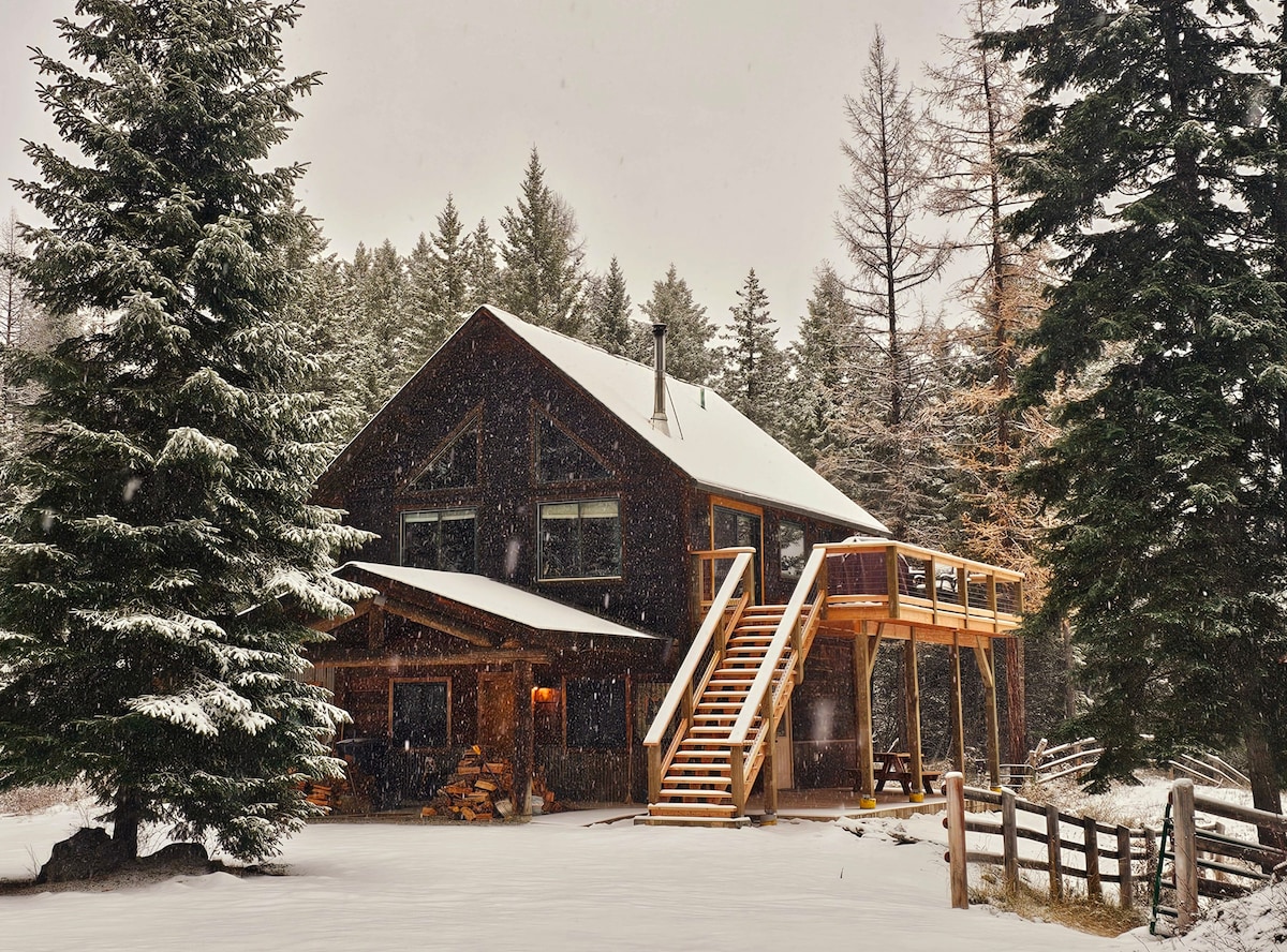 A cozy cabin nestled among tall evergreen trees is blanketed in fresh snow. A wooden deck with stairs leads up to the entrance, inviting guests into a warm retreat. Snowflakes gently fall, adding a serene winter ambiance to the rustic exterior.