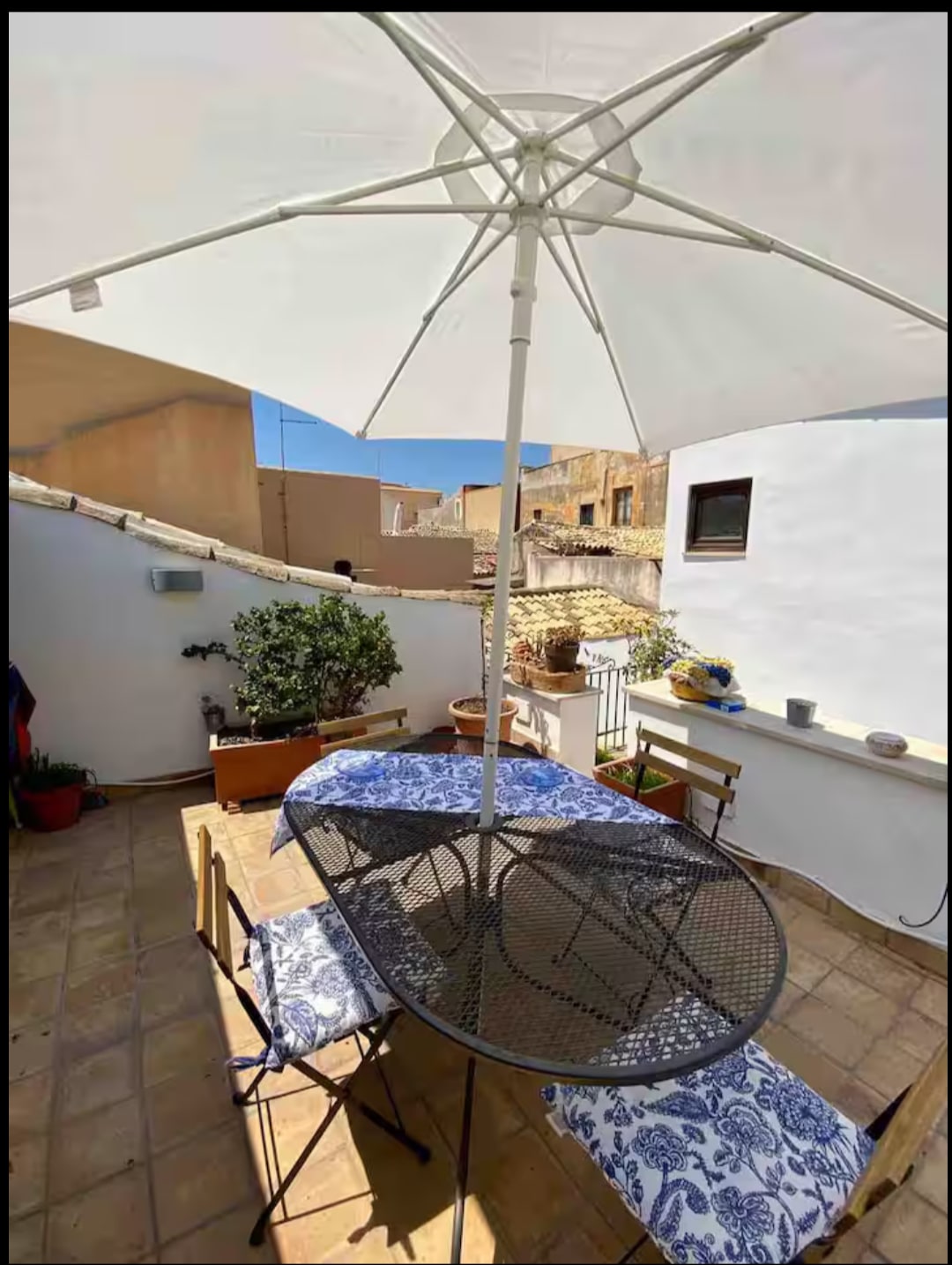 A shaded outdoor terrace features a round table surrounded by two chairs, with a blue and white patterned tablecloth. Potted plants enhance the space, while a small wall serves as a backdrop. Sunlight brightens the area, creating a pleasant setting for relaxation.