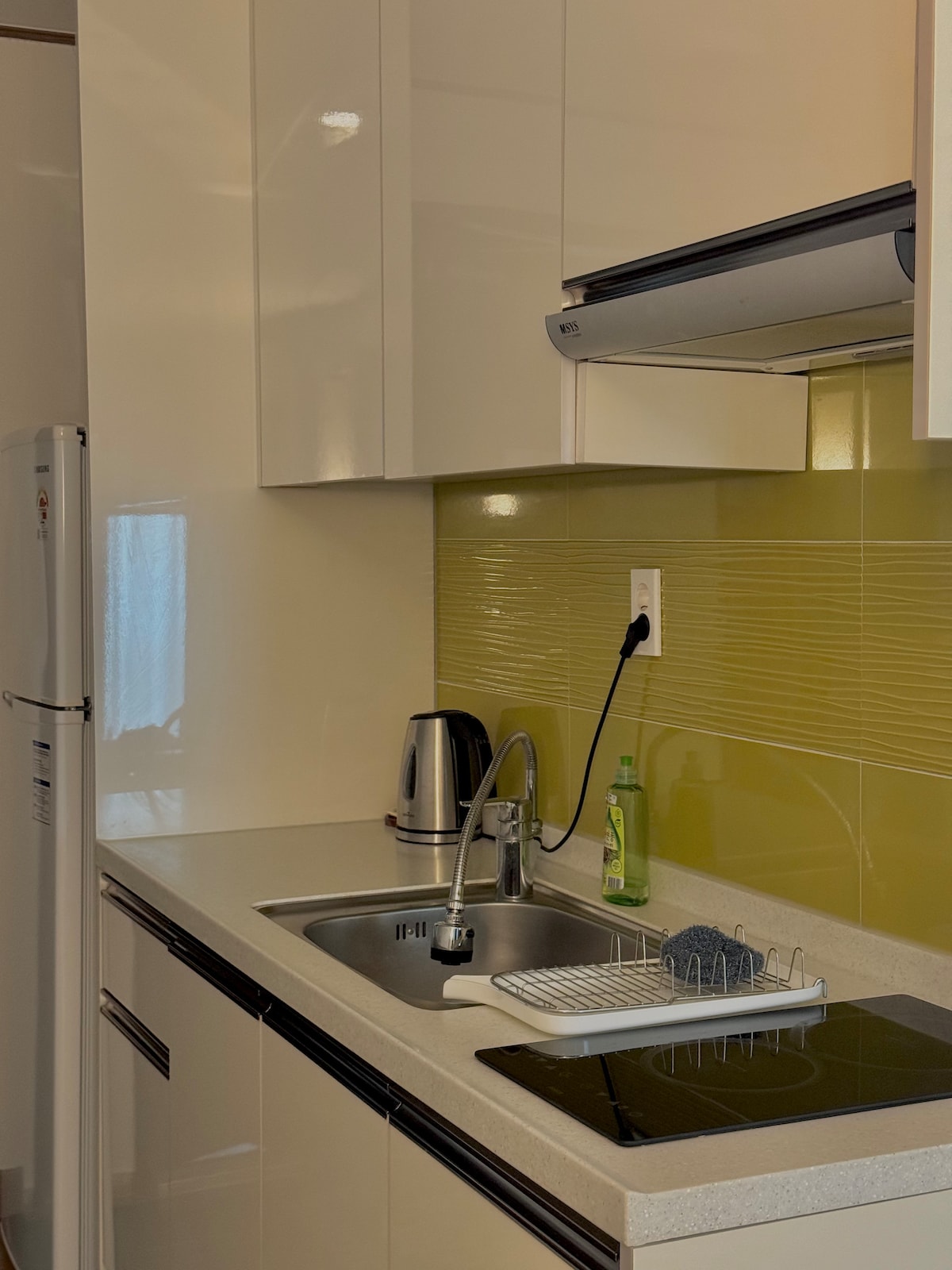 A modern kitchen space features sleek white cabinetry and a bright green backsplash. A stainless steel kettle sits next to a sink, with a dish drying rack and an induction cooktop on the counter. Natural light filters through a nearby window.