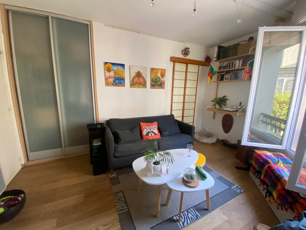 A cozy living area features a dark sofa adorned with a decorative cushion. A round table is set in front, accompanied by a small plant. Natural light floods in from a large window, illuminating the shelves that display books and decorative items.