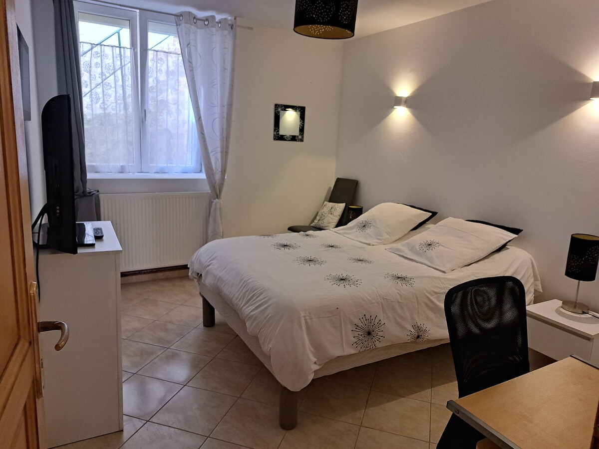 A well-lit bedroom features a spacious bed adorned with a decorative cover. Two black bedside lamps offer subtle lighting, while a desk and chair provide a workspace. Natural light enters through large windows with sheer curtains, creating a serene atmosphere.