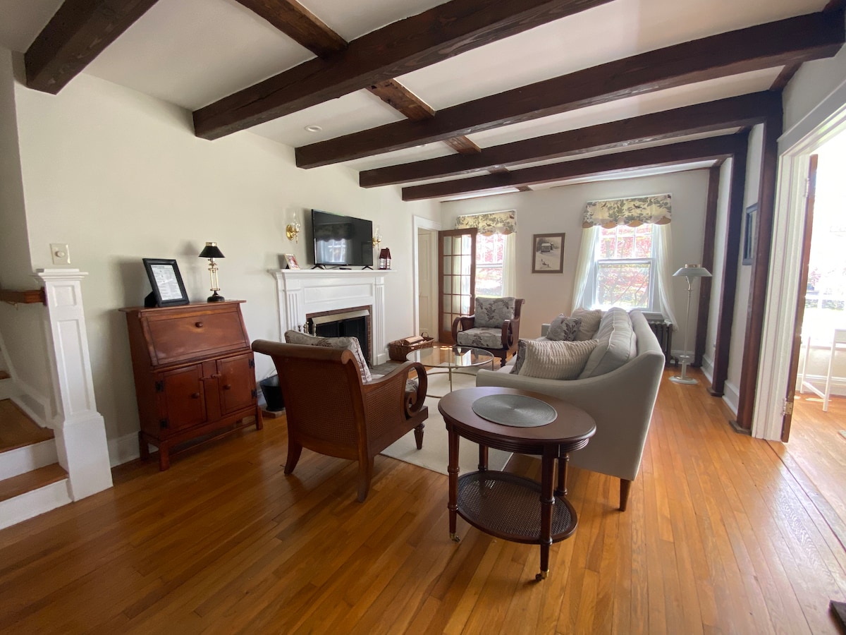 The living room features original beams and hardwood floors, with a cozy arrangement of comfortable seating. A smart TV is mounted above a classic fireplace, while sunlight filters through multiple windows, creating a warm ambiance. A small antique desk is also visible, adding character.