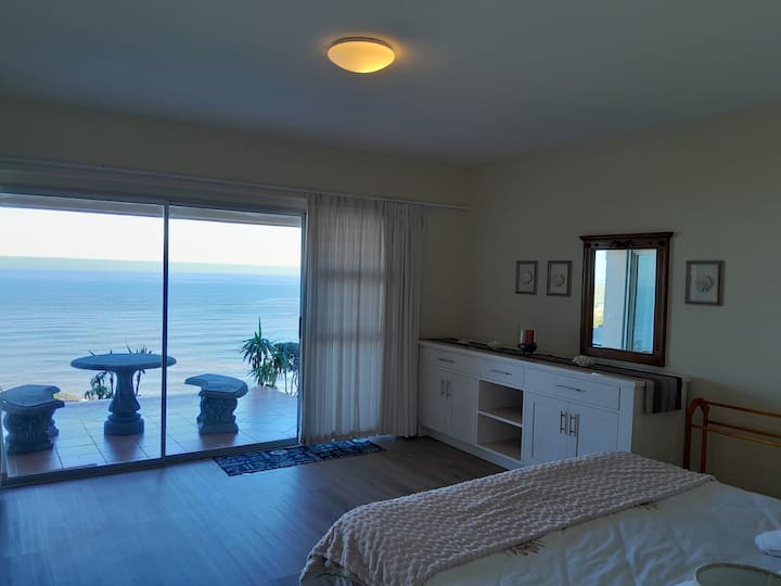 Pamje e oqeanit me karrige dhe tavolina guri jashtë derës.