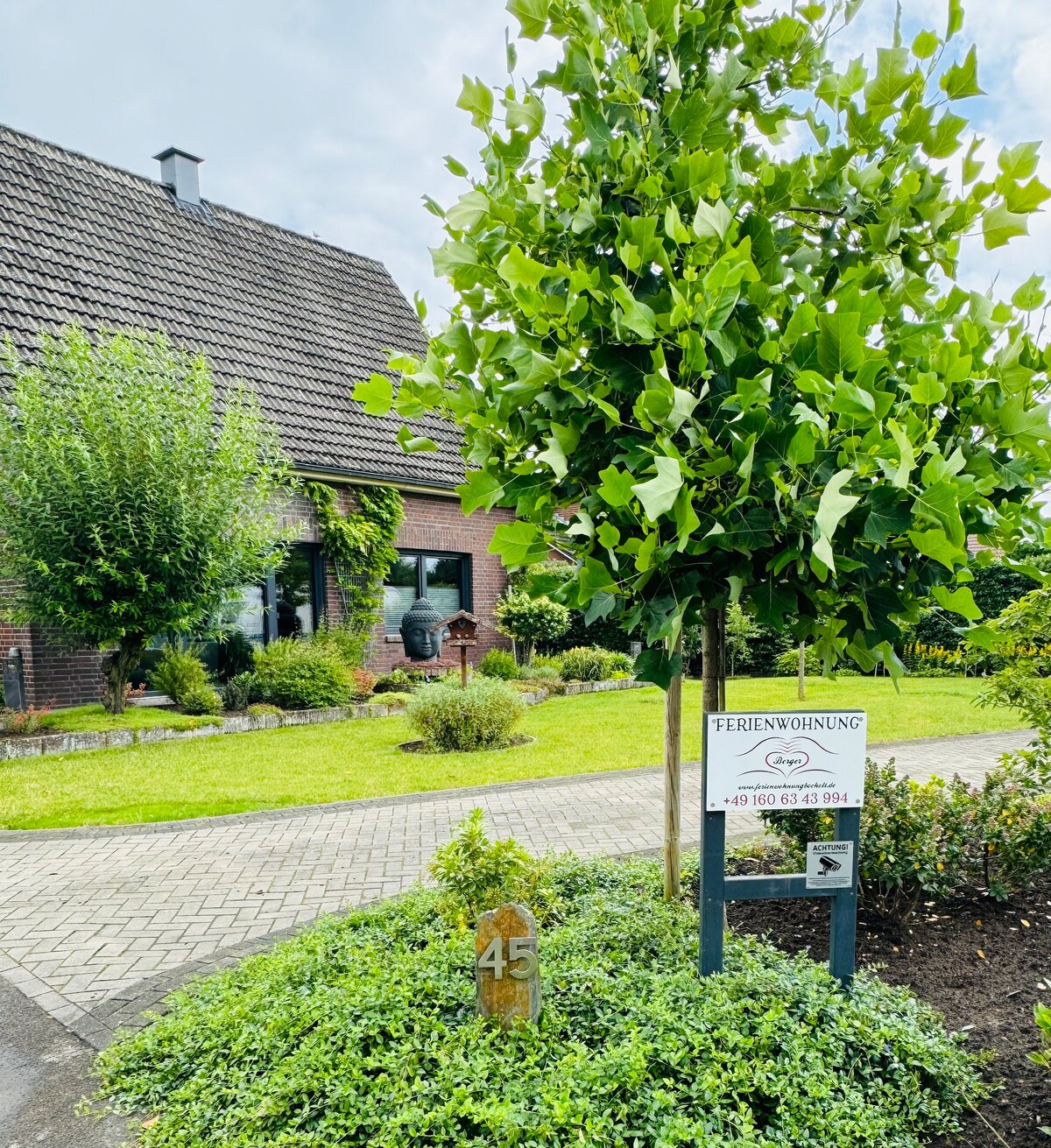 A welcoming exterior of the property is displayed, showcasing a well-maintained garden with various shrubs and trees. A sign indicating 'Ferienwohnung' is prominently featured, along with a house number. The charming facade includes a mix of brick and greenery, offering an inviting atmosphere.