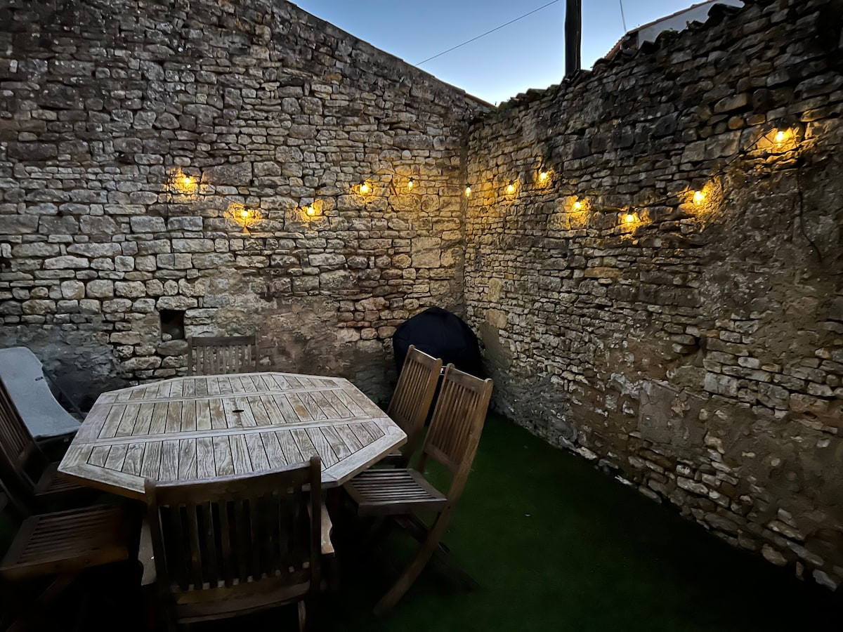 An outdoor dining area features a round wooden table surrounded by six wooden chairs, illuminated by soft string lights. The rustic stone walls add character to the space, while a dark blue object, possibly a cover, is positioned in the corner.