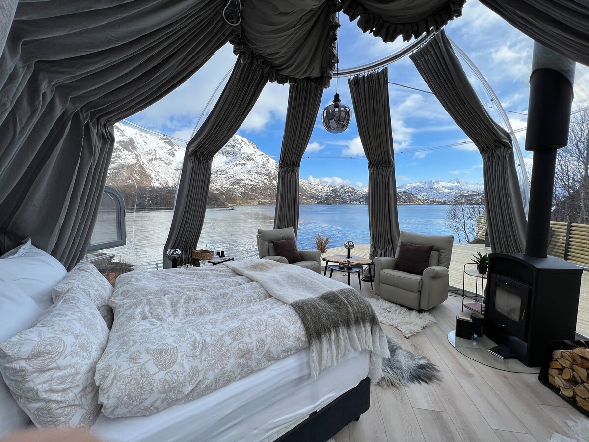 A cozy sleeping area features a spacious bed with soft bedding and a warm throw. Comfortable seating faces large windows, offering a stunning view of the water and mountains. A stylish wood stove adds ambiance to the space, enhancing the connection with nature outside.