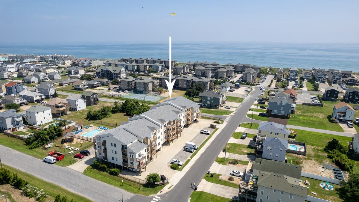An aerial view of Sundune Village is displayed, illustrating the three-story buildings situated near the coastline. The community pool can be seen in the distance, while the surrounding areas feature residential homes and open spaces. The ocean is visible beyond the neighborhood.