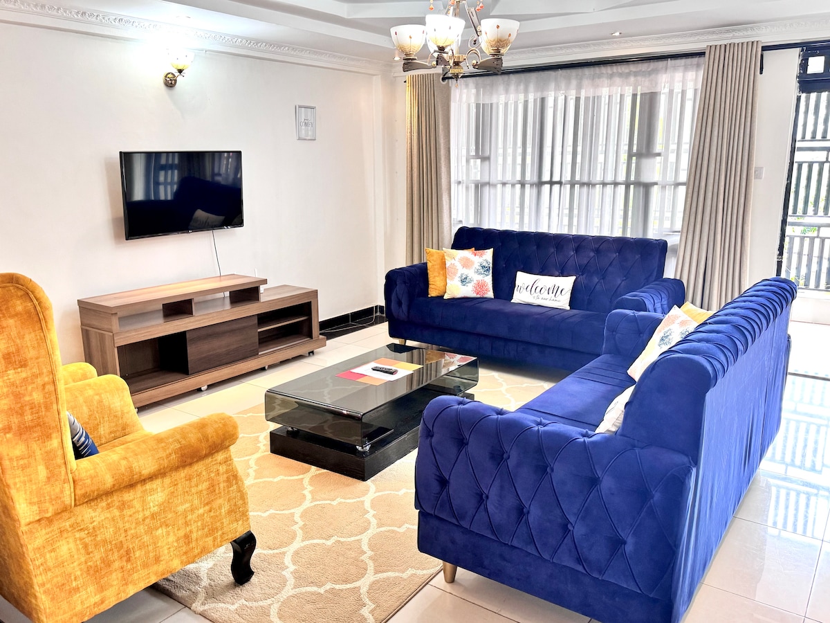 The spacious living area features a bright blue sofa set and a cozy yellow armchair, arranged around a black coffee table. A flat-screen TV is mounted on the wall, and large windows provide natural light, complemented by light-colored curtains.