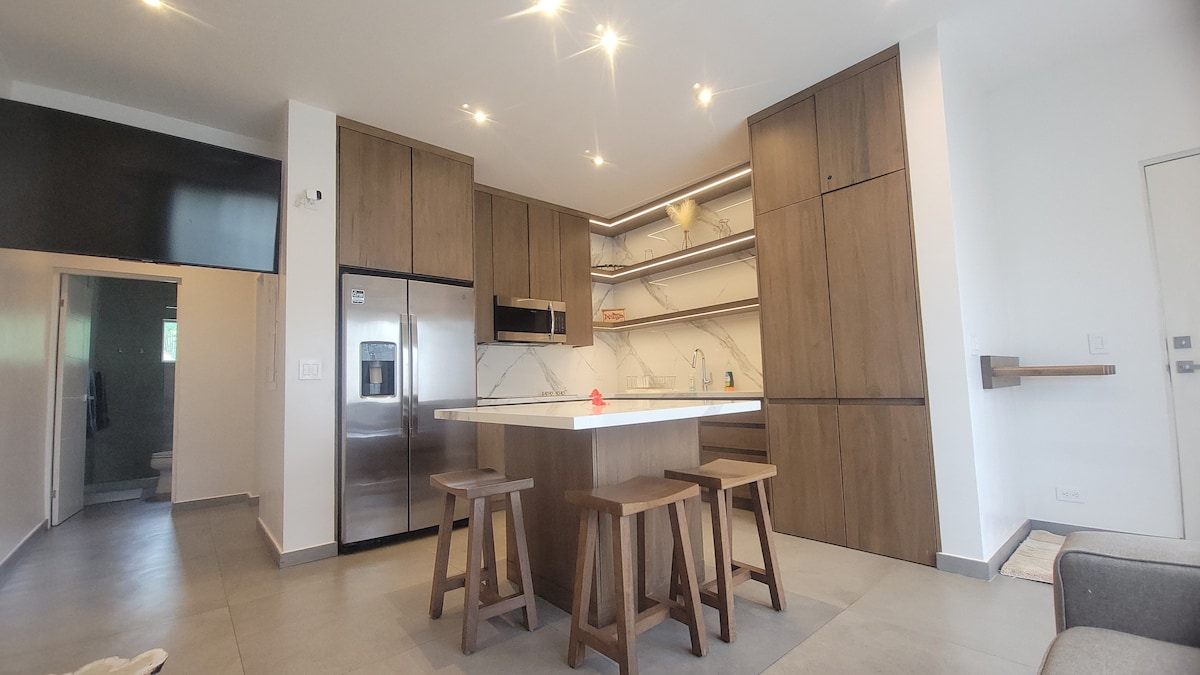 An open kitchen area features modern cabinetry and stainless steel appliances. A marble-topped island with three wooden stools is positioned at the center. Shelving provides storage above, and soft lighting creates a bright atmosphere. The layout connects seamlessly to the living space.