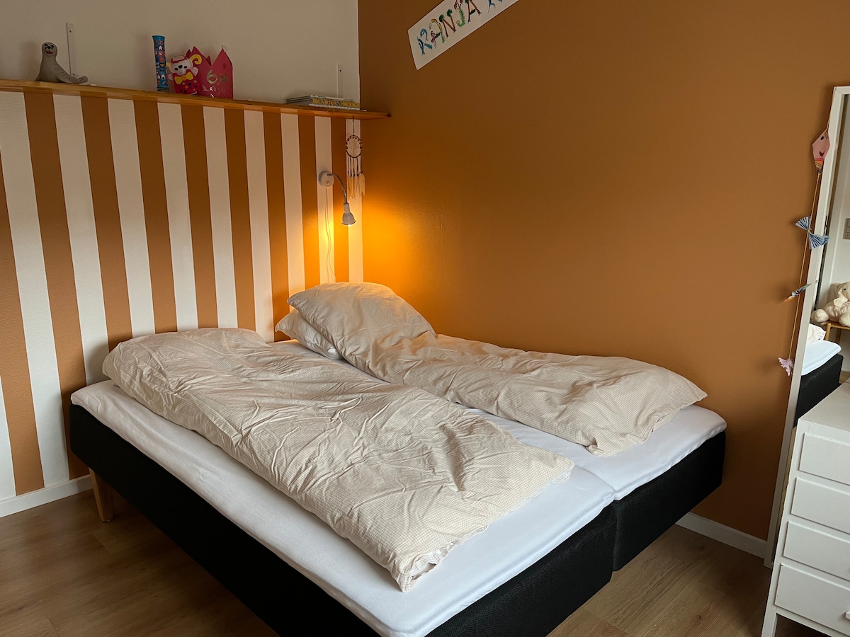 A bedroom features two twin beds, situated against a warm, brown accent wall with cream and white striped decor. A bedside lamp provides soft illumination, while a simple dresser is positioned nearby. Shelves hold decorative items, adding a personal touch to the space.