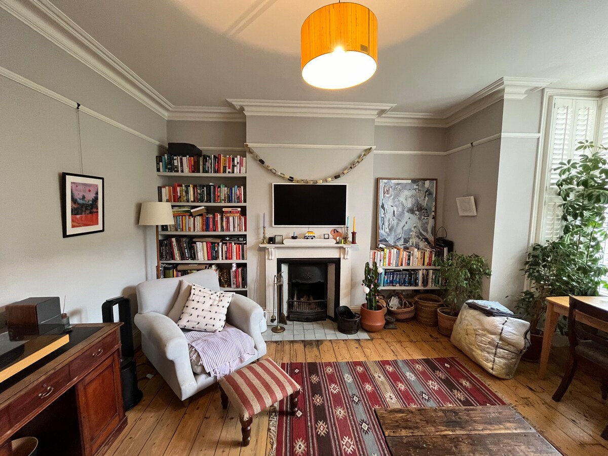 A spacious living room features a cozy armchair and a wooden table. A fireplace is centered against the wall, with a television mounted above it. Bookshelves filled with books line one side, and potted plants add greenery to the room. Wooden flooring provides a warm feel.
