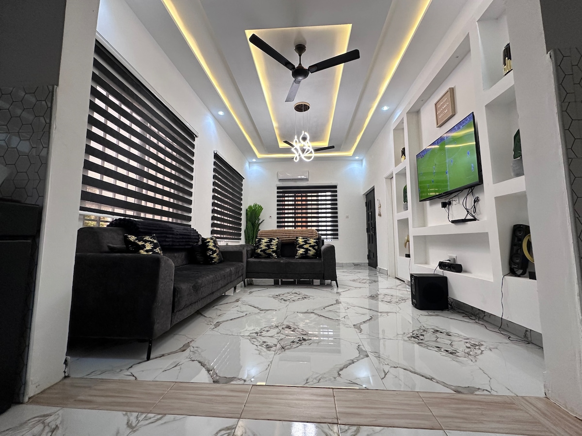 A spacious living area features modern decor with a dark sectional sofa and decorative cushions. Bright overhead lighting complements the clean marble floor, while striped window treatments control natural light. A wall-mounted television is visible, alongside built-in shelves that hold various audio equipment.