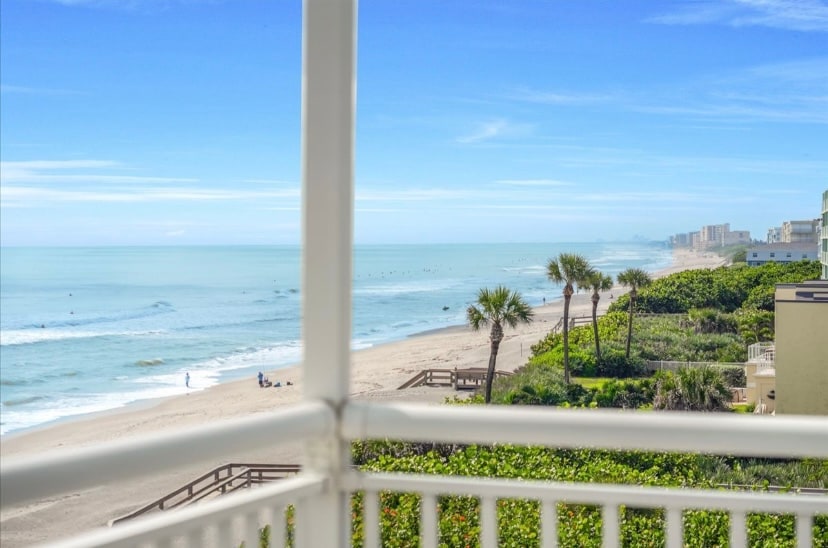 A stunning ocean view is displayed from the balcony, featuring gentle waves lapping against the sandy beach. Palm trees line the shore, and the distant coastline is visible under a clear blue sky.