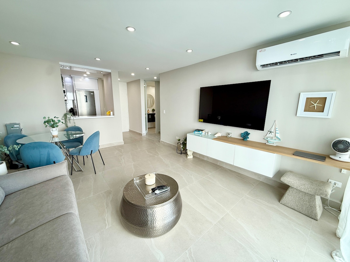 The living area features a neutral color palette with a gray sofa facing a large wall-mounted television. A round coffee table sits in the center, complemented by blue dining chairs and a view of the kitchen area. Bright lighting enhances the open, spacious feel.
