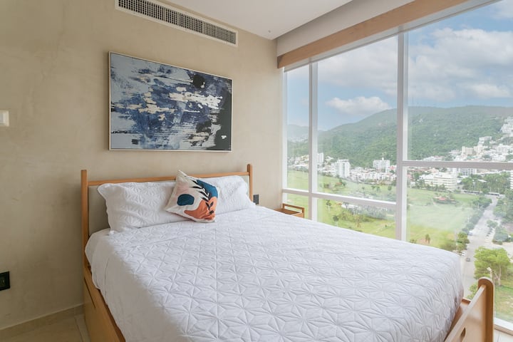 Cama queen junto a un ventanal con hermosa vista, decoración en tonos suaves y un ambiente lleno de luz natural.