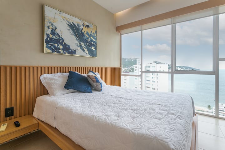 Cama queen con luz natural y un cuadro en tonos azules que crea un ambiente relajante y moderno.