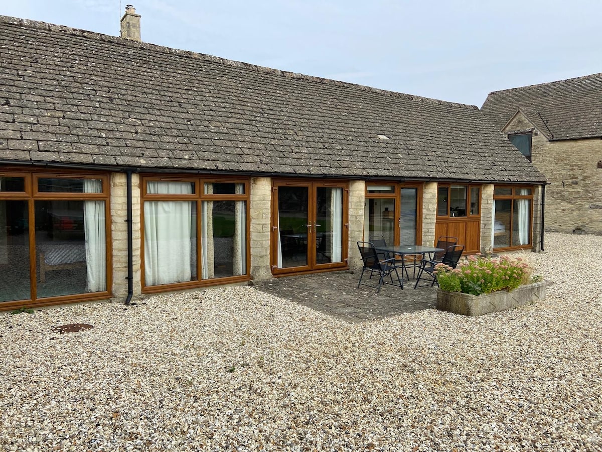 The exterior of the cottage showcases a single-storey structure with large, south-facing windows and French doors, providing access to a patio area. A small round table with chairs is positioned on a gravel surface surrounded by low stone borders filled with greenery.