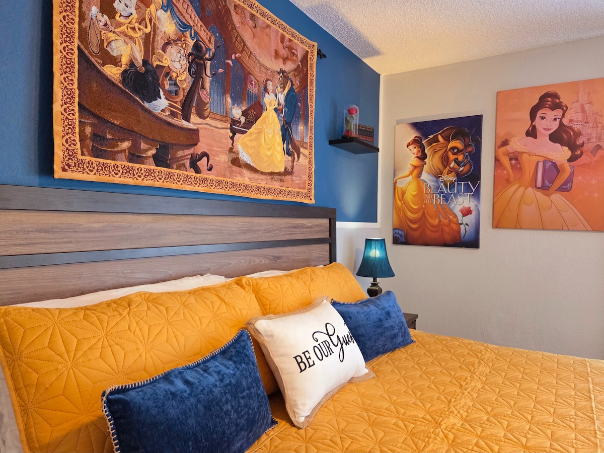A cozy bedroom features a large bed adorned with an orange quilt and decorative pillows. Colorful wall art depicting fairytale characters is displayed above the bed, set against a blue backdrop. A bedside table holds a green lamp, contributing to the inviting atmosphere.