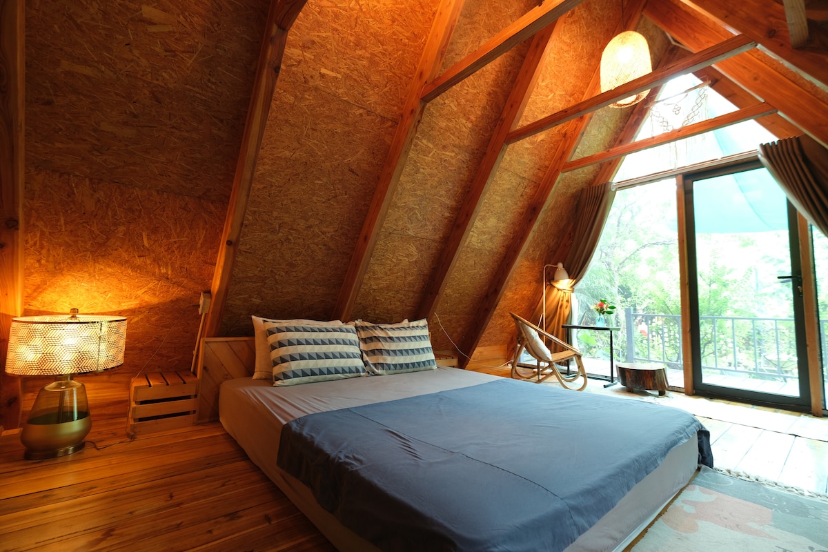 An open, attic-style bedroom showcases a spacious bed with neatly arranged pillows. Natural wood textures and warm lighting create a cozy ambiance. A large window offers views of greenery outside, while a seating area with a wooden accent chair adds to the inviting space.