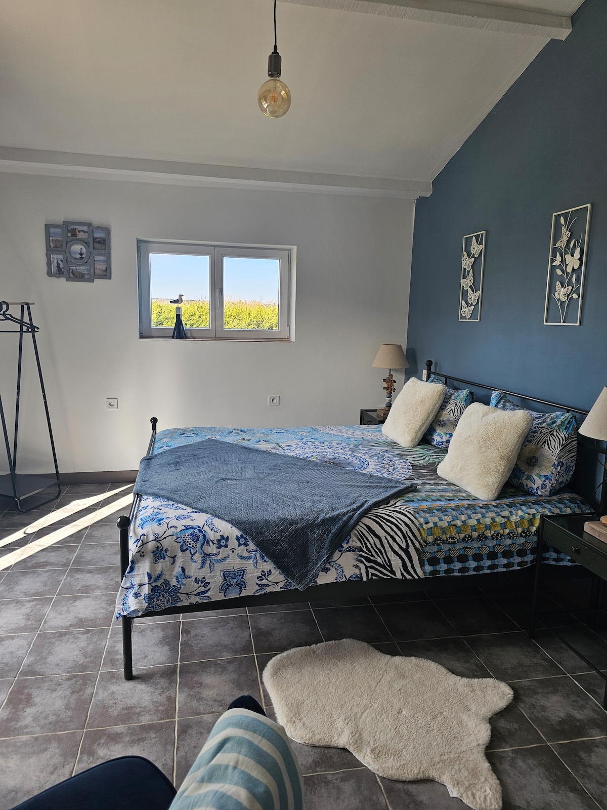 A well-lit bedroom features a spacious bed adorned with a colorful patchwork quilt and decorative cushions. A window provides natural light, while artwork and lamps add charm. A cozy rug lies on the tiled floor, and a clothing rack offers additional storage.