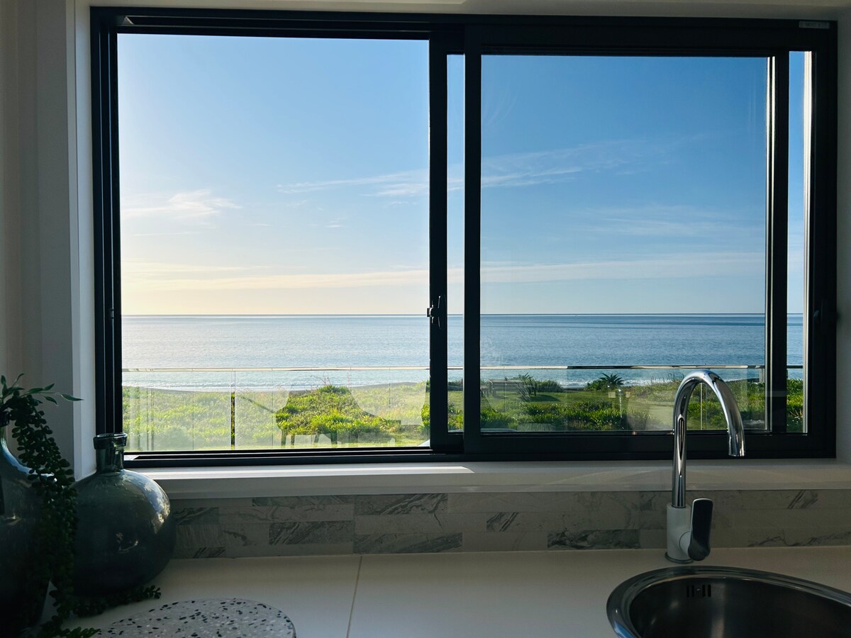 A large window frames a serene view of the ocean, showcasing calm blue waters and a clear sky. The kitchen counter is partially visible, highlighting modern fixtures and decorative elements such as plants and vases.