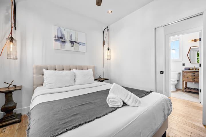 Bright and inviting guest bedroom featuring crisp white linens, dark gray throw accents, and modern wall lighting.