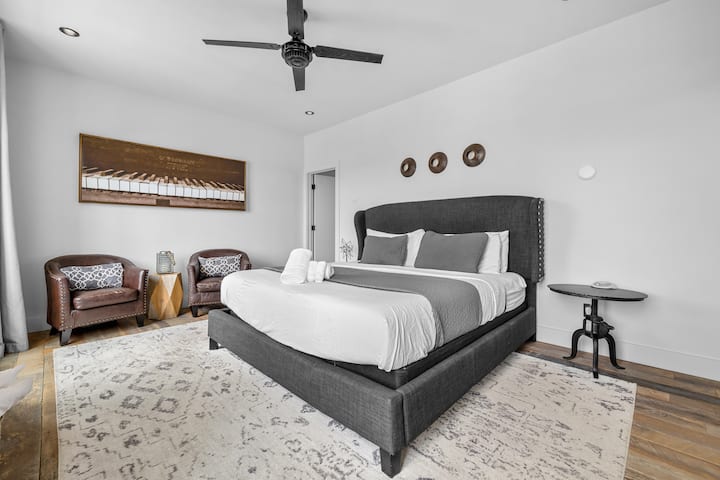Spacious primary bedroom featuring a dark upholstered king bed, minimalist white walls, and a modern ceiling fan.