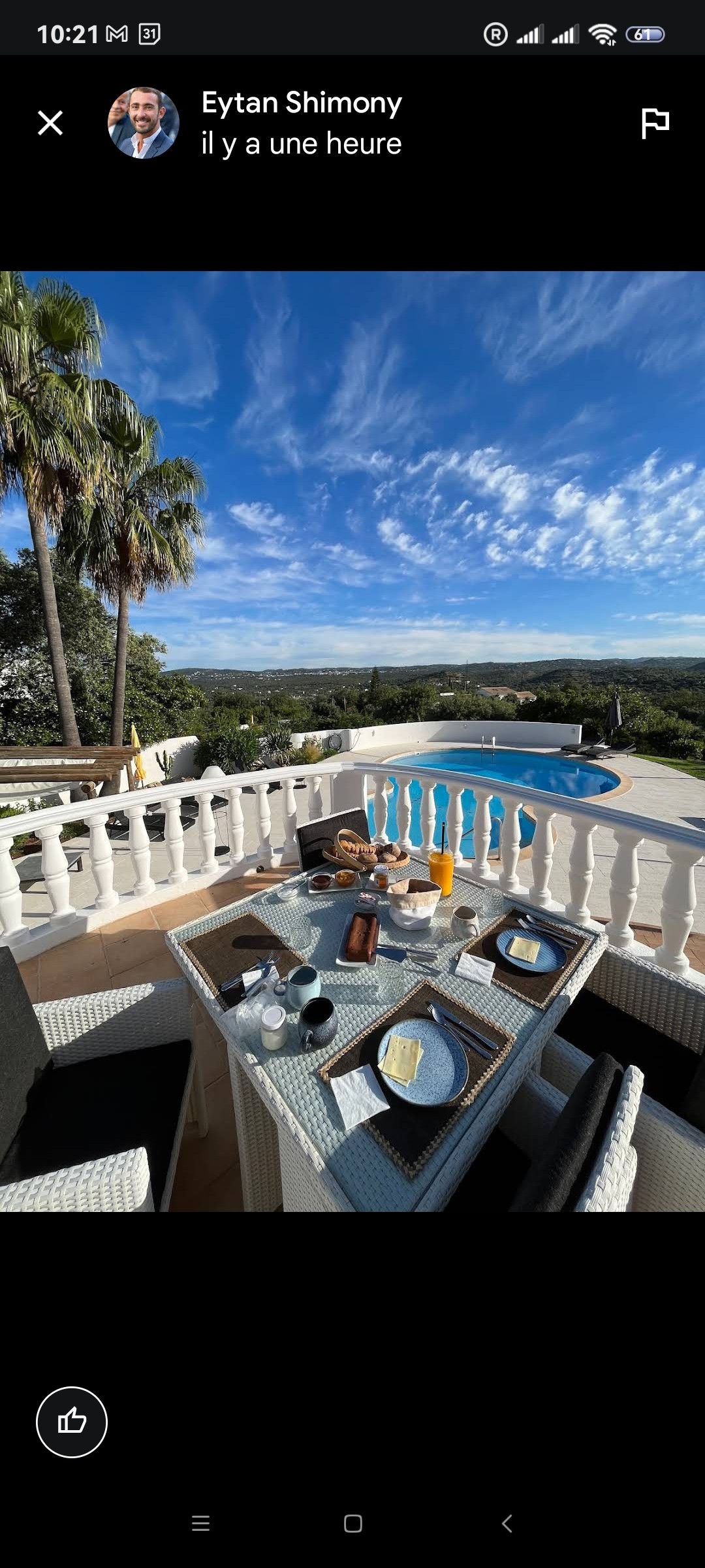 A private terrace features a dining table set for breakfast with plates and drinks, overlooking a serene view of rolling hills and a pool. Surrounding greenery and palm trees complement the tranquil ambiance, with a clear blue sky above.