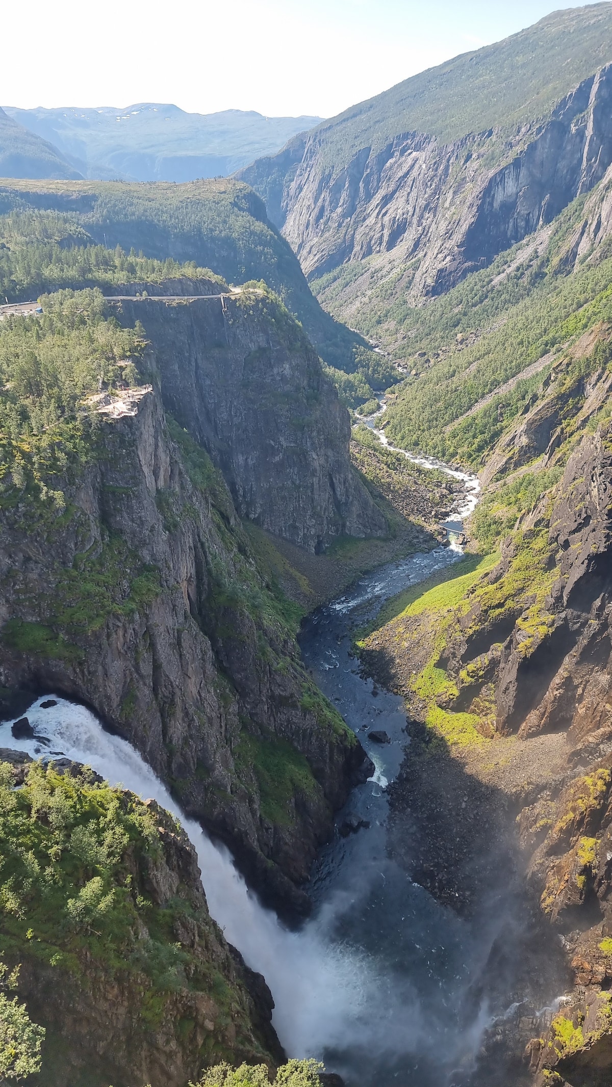 A panoramic view of a steep, rocky canyon is presented, with a waterfall cascading into a river below. Lush green vegetation is visible along the canyon walls, contrasting with the rugged terrain. The distant mountains provide a dramatic backdrop against a clear sky.