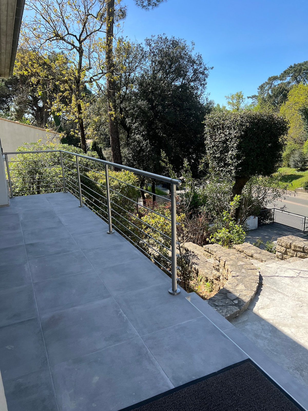 A spacious terrace features modern railing and grey tiled flooring, surrounded by lush greenery and trees. The view extends to a pathway and landscaped garden, providing a serene outdoor space for relaxation.
