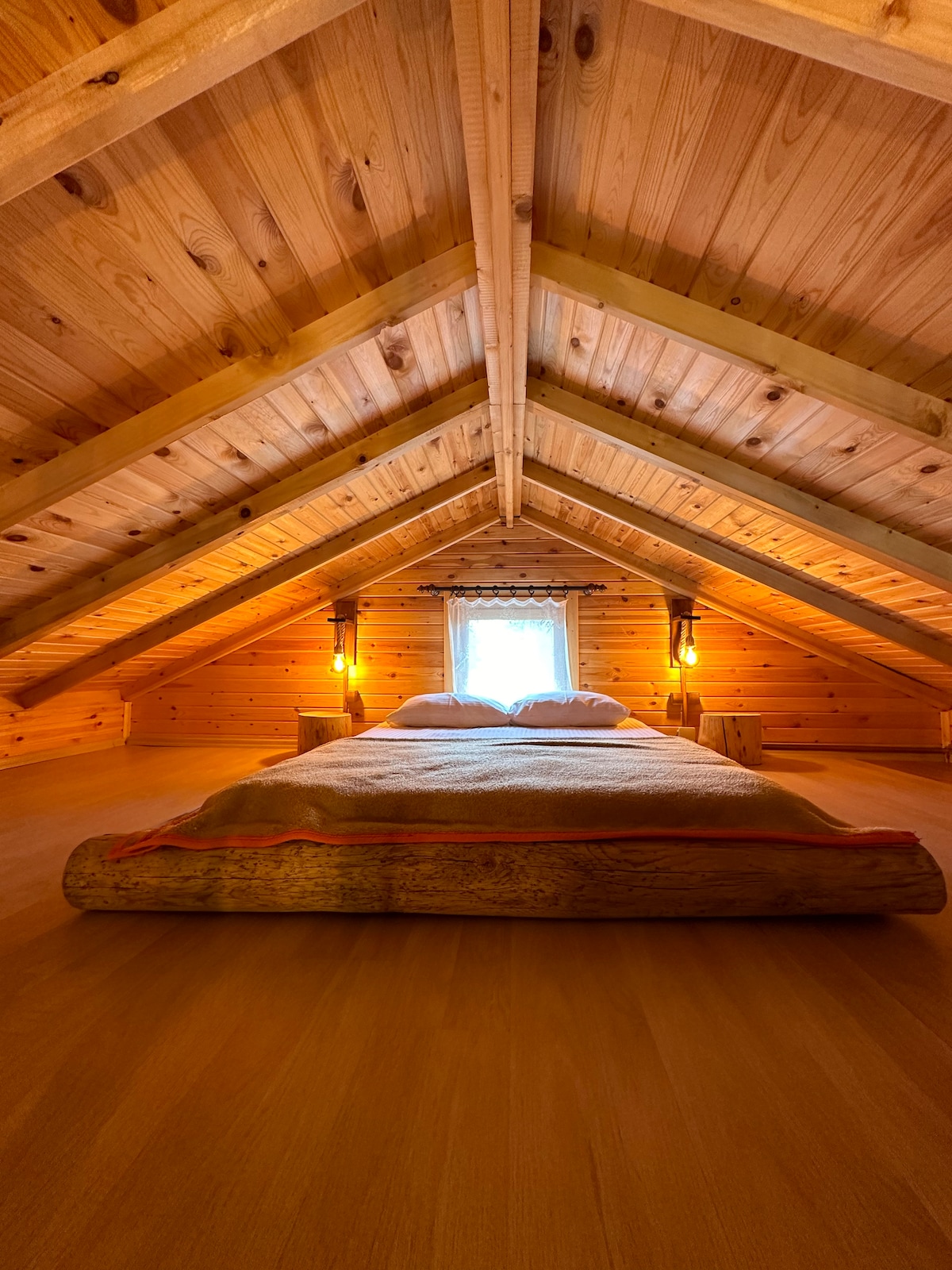 A spacious sleeping area is situated under a sloped wooden ceiling, featuring warm-toned wooden walls. Soft lighting from wall sconces provides a gentle glow, while a cozy bed is positioned at the center, with light filtering through a small window adorned with a lace curtain.