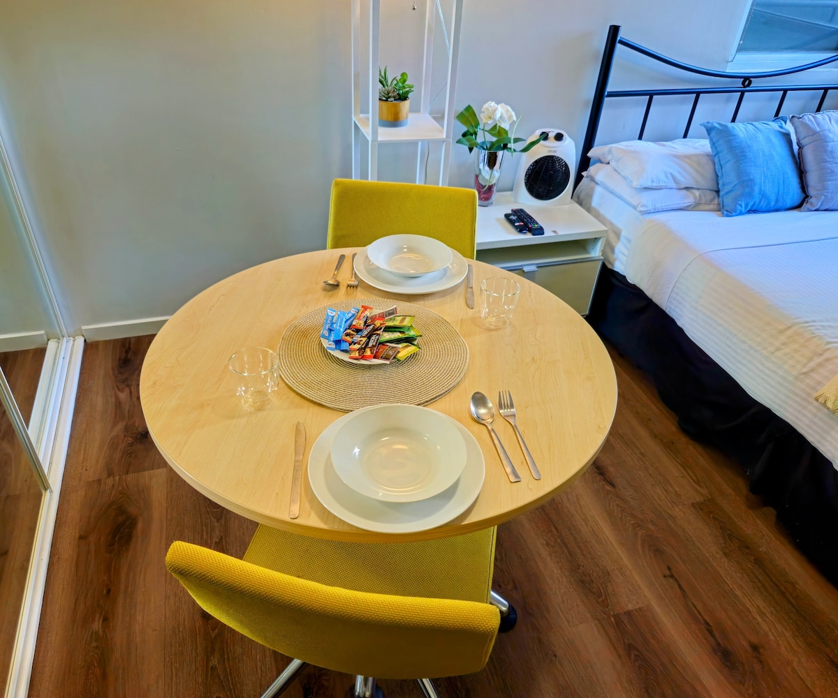 A round wooden dining table is set for two, featuring white plates and glassware. A colorful platter of appetizers is displayed at the center. Yellow chairs are positioned around the table, with soft lighting creating a comfortable dining area adjacent to the sleeping space.