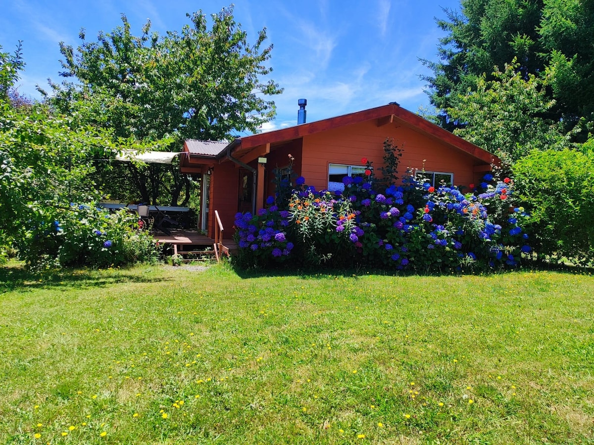 A charming wooden cottage is surrounded by vibrant flowers and greenery. The garden features colorful blooms, including hydrangeas, and a seating area on the porch. The clear skies above complement the peaceful rural setting, providing a serene atmosphere for relaxation.