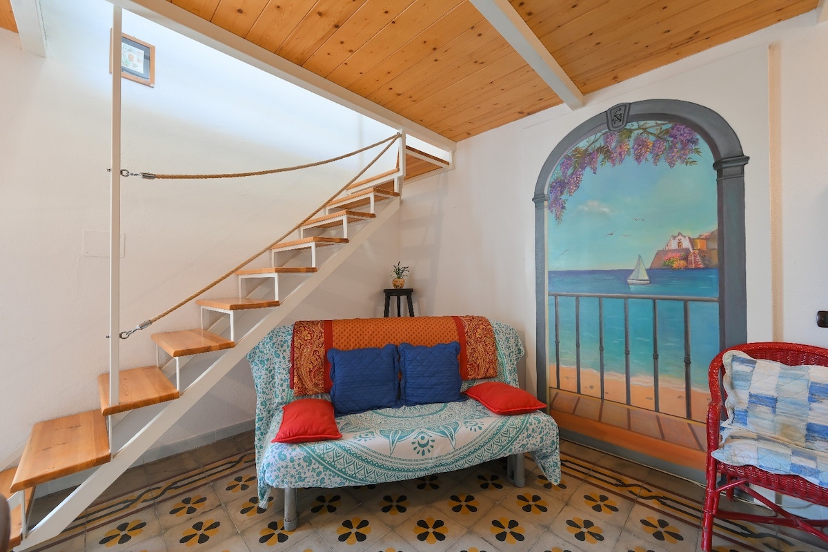 A seating area featuring a colorful couch adorned with decorative cushions is complemented by a mural depicting a coastal view. An open staircase with wooden steps leads to the upper level, while the tiled floor adds character to the space.