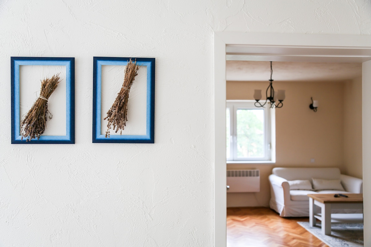 Two framed bundles of dried herbs hang on a light-colored wall near an open doorway. A cozy seating area is visible in the adjacent room, featuring a light-colored sofa and a wooden coffee table, creating a relaxed atmosphere.