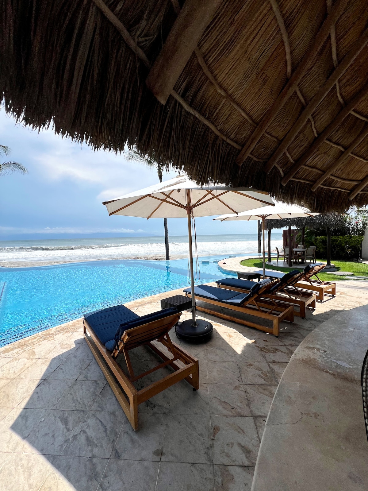 Lounge chairs with blue cushions are positioned beside a shimmering infinity pool, providing a view of the ocean. Large umbrellas offer shade, while a thatched roof adds a tropical touch to the setting. The surrounding area features lush greenery and well-maintained landscaping.