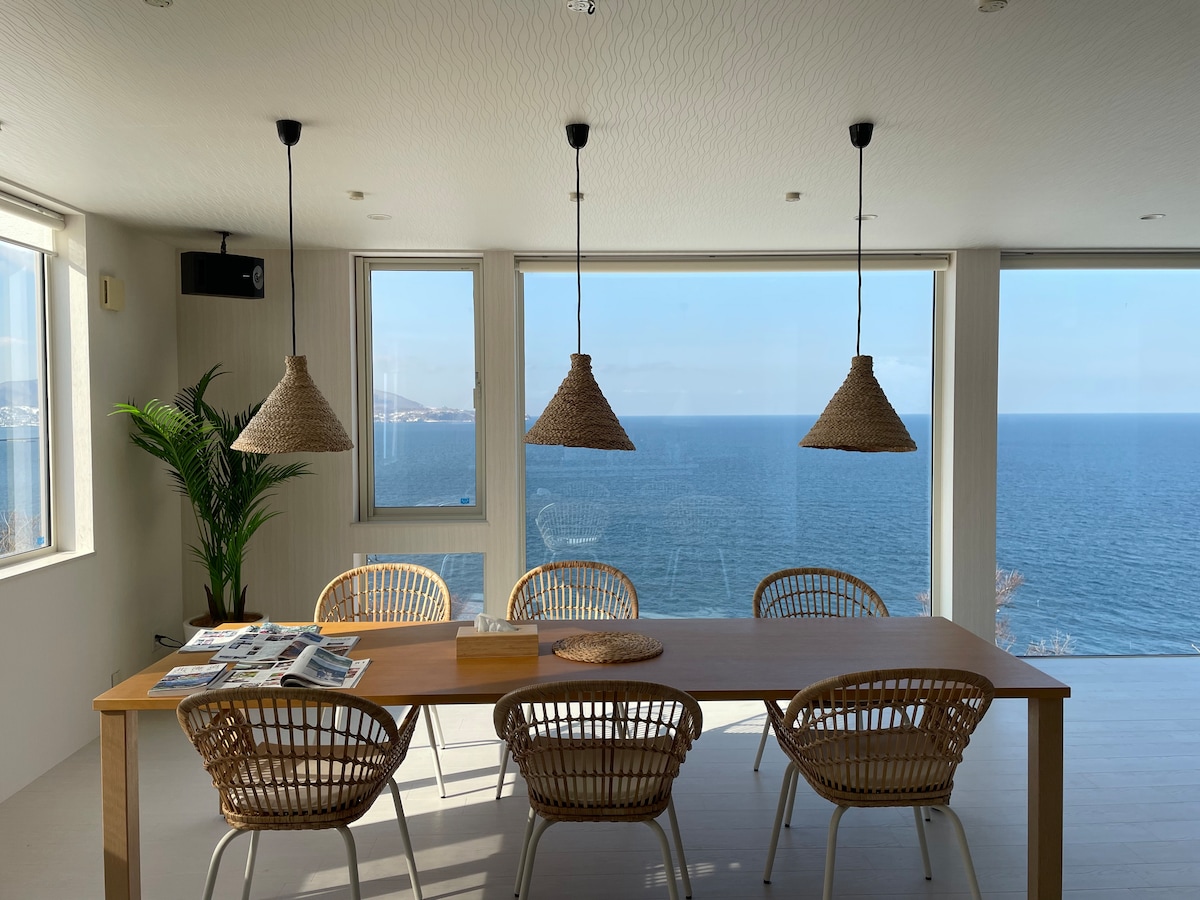 A dining area is presented with a wooden table surrounded by six wicker chairs. Three pendant lights hang above, illuminating the space. Large windows offer an unobstructed view of the sea, allowing natural light to enhance the bright interior.