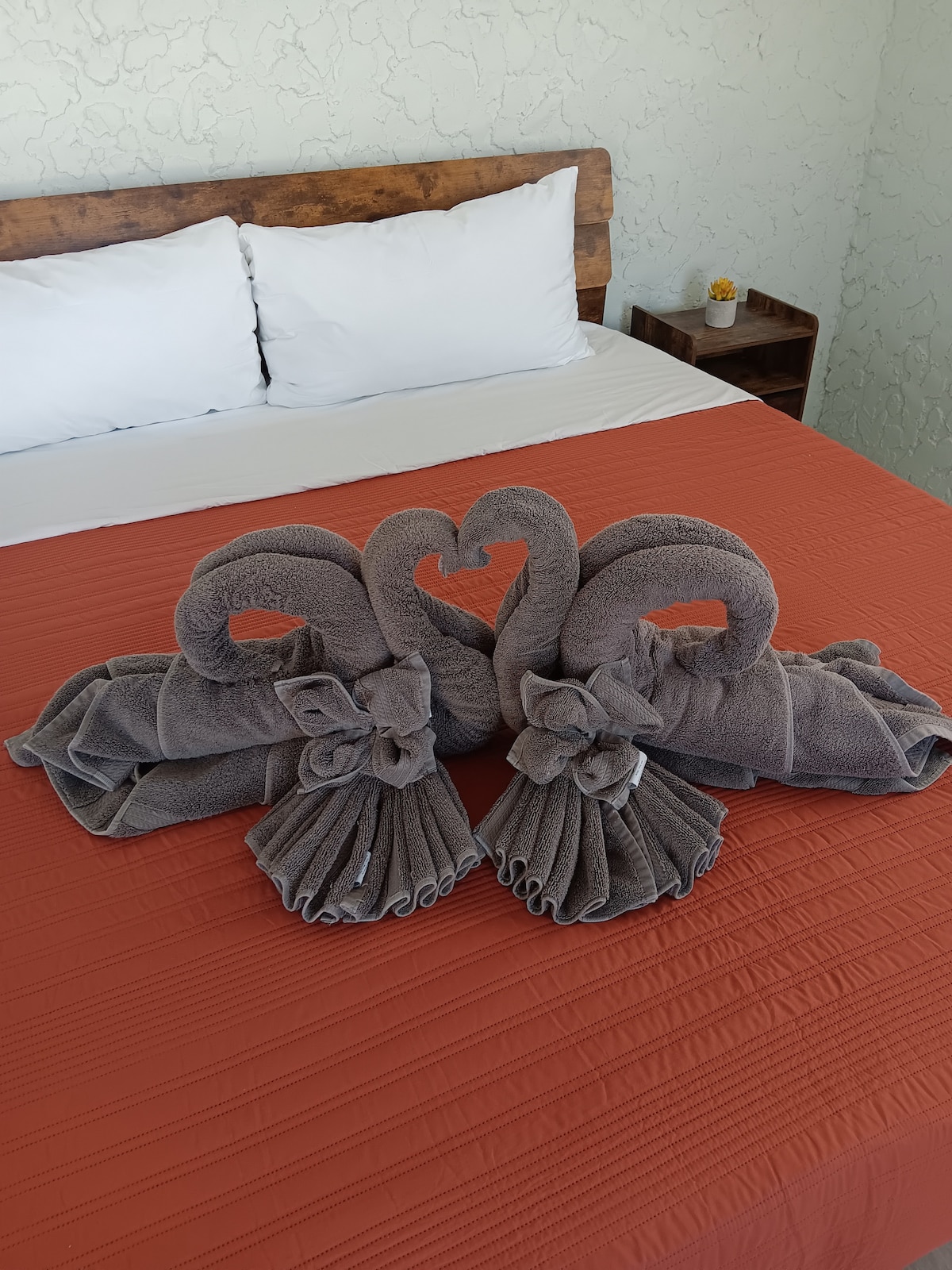 A spacious bed is dressed in an orange coverlet, featuring white pillows at the headboard. Two decorative swans crafted from gray towels are arranged on the bed, creating a heart shape with their necks, adding a unique touch to the room.