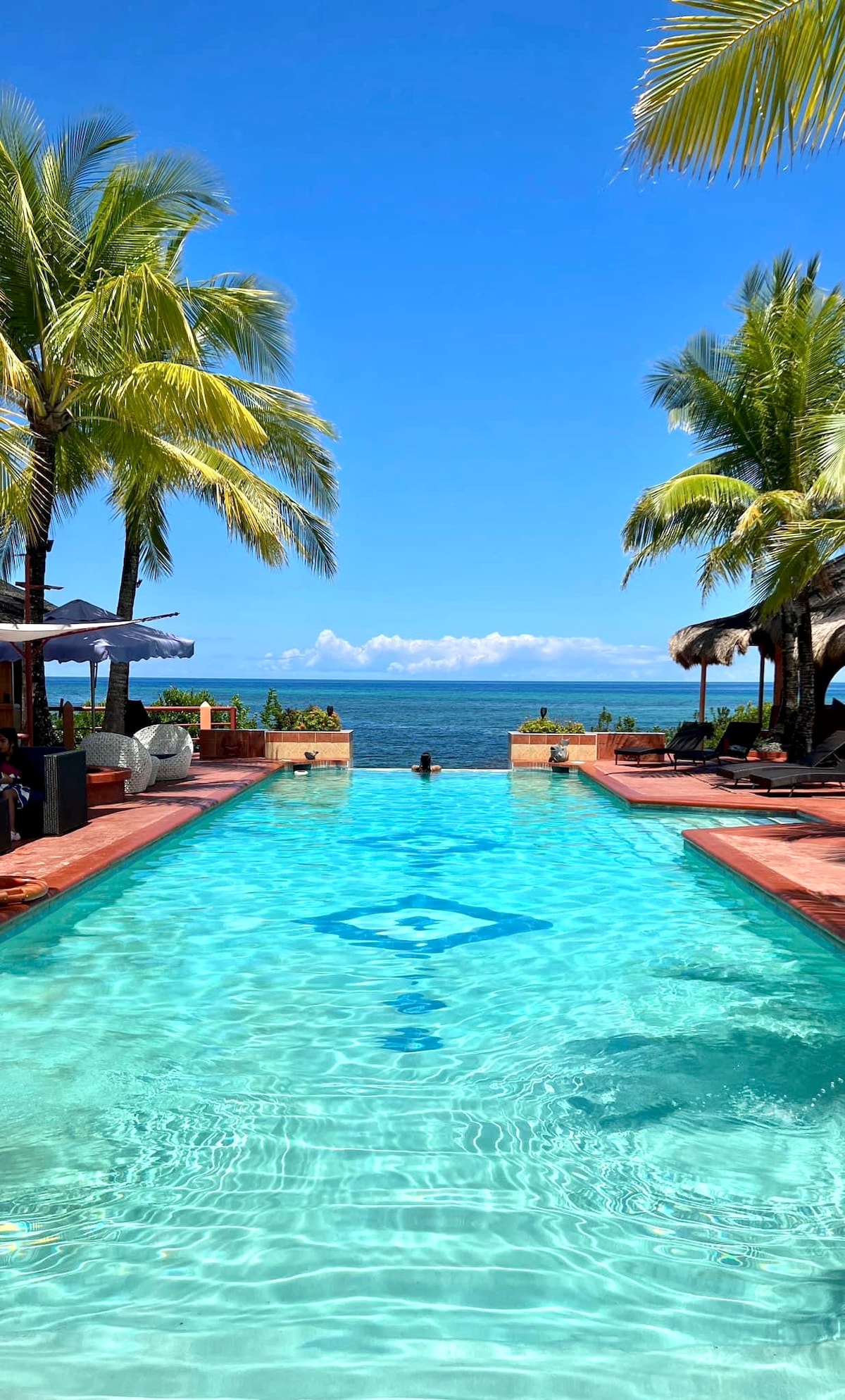 A clear infinity pool extends towards the horizon, reflecting the vibrant blue sky. Lush palm trees are seen on either side, providing shade. Comfortable lounge chairs are positioned around the pool area, offering a place to relax and enjoy the view of the sea.
