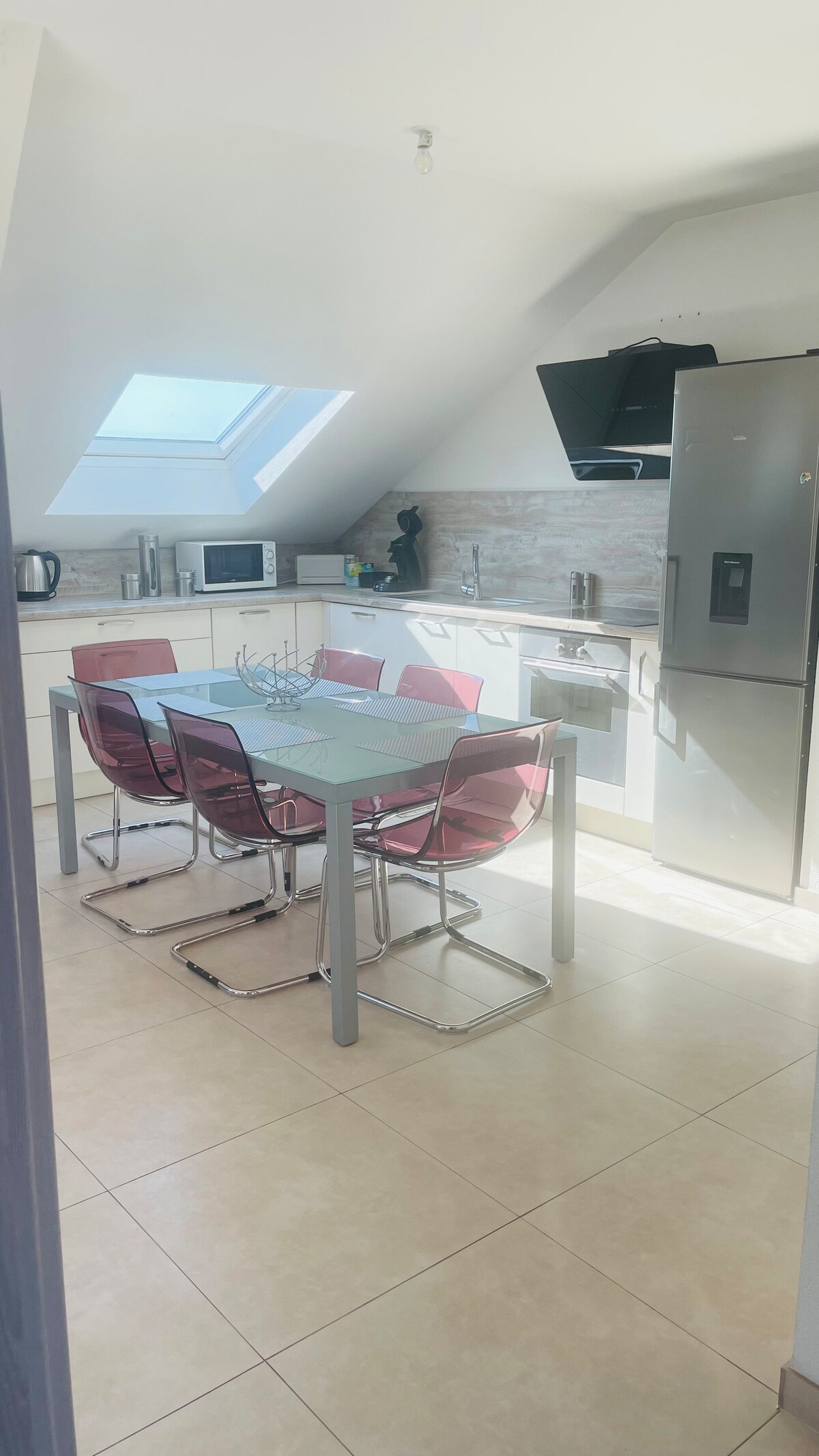 A modern kitchen is visible, featuring a light-colored countertop and ample cabinetry. A glass dining table is surrounded by six transparent chairs. Natural light filters in through a skylight, illuminating the space. Appliances like a refrigerator and microwave are positioned conveniently.