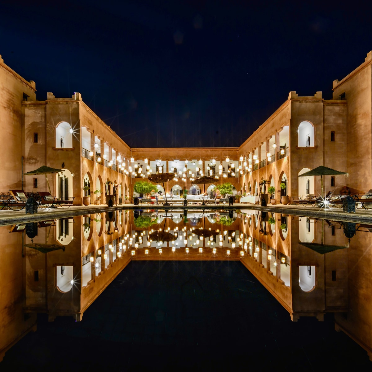 The kasbah is illuminated at night, reflecting soft lights in the dark water of the central pool. Arched doorways and lanterns create a serene atmosphere, while plants and seating areas enhance the inviting communal space around the water.
