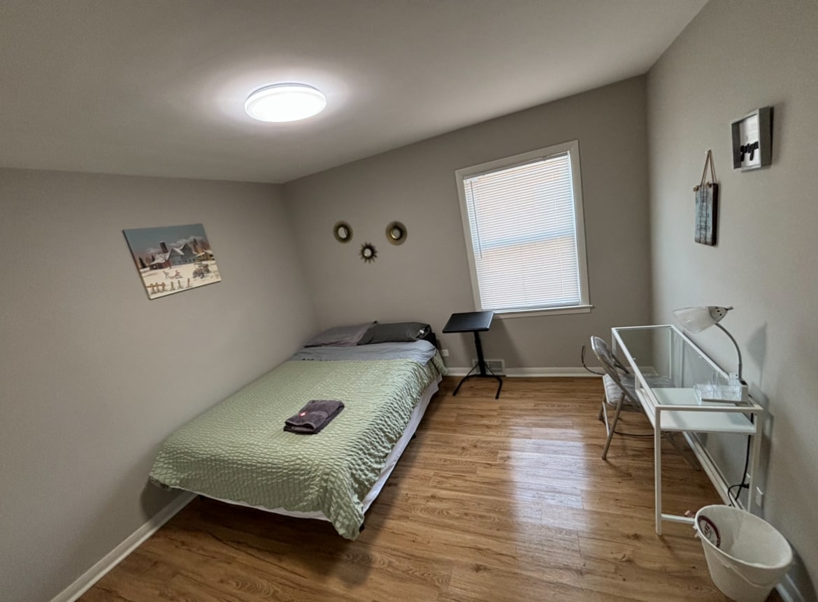 A simple bedroom features a spacious bed dressed with a mint-colored blanket. A small desk and chair are positioned next to a window, with natural light filtering through. Wall decor includes mild artwork and circular mirrors, complementing the wood-style flooring.