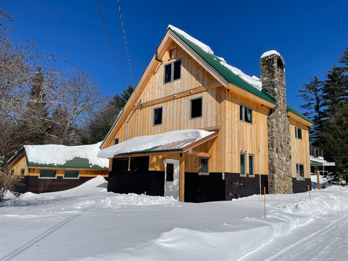The exterior of a mountain lodge is framed by a blanket of fresh snow. The wooden structure features a steeply pitched roof, a prominent stone chimney, and large windows. The entrance is covered by a small awning, while the surrounding landscape includes snow-draped trees.