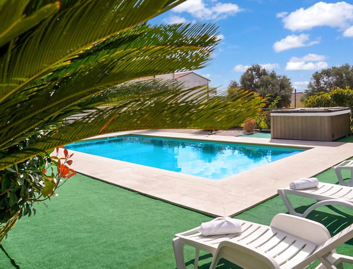 An outdoor pool is framed by a manicured green lawn, providing space for relaxation. White lounge chairs are positioned nearby, each featuring a neatly folded towel. Lush greenery and colorful plants are visible around the pool area, contributing to a serene environment.