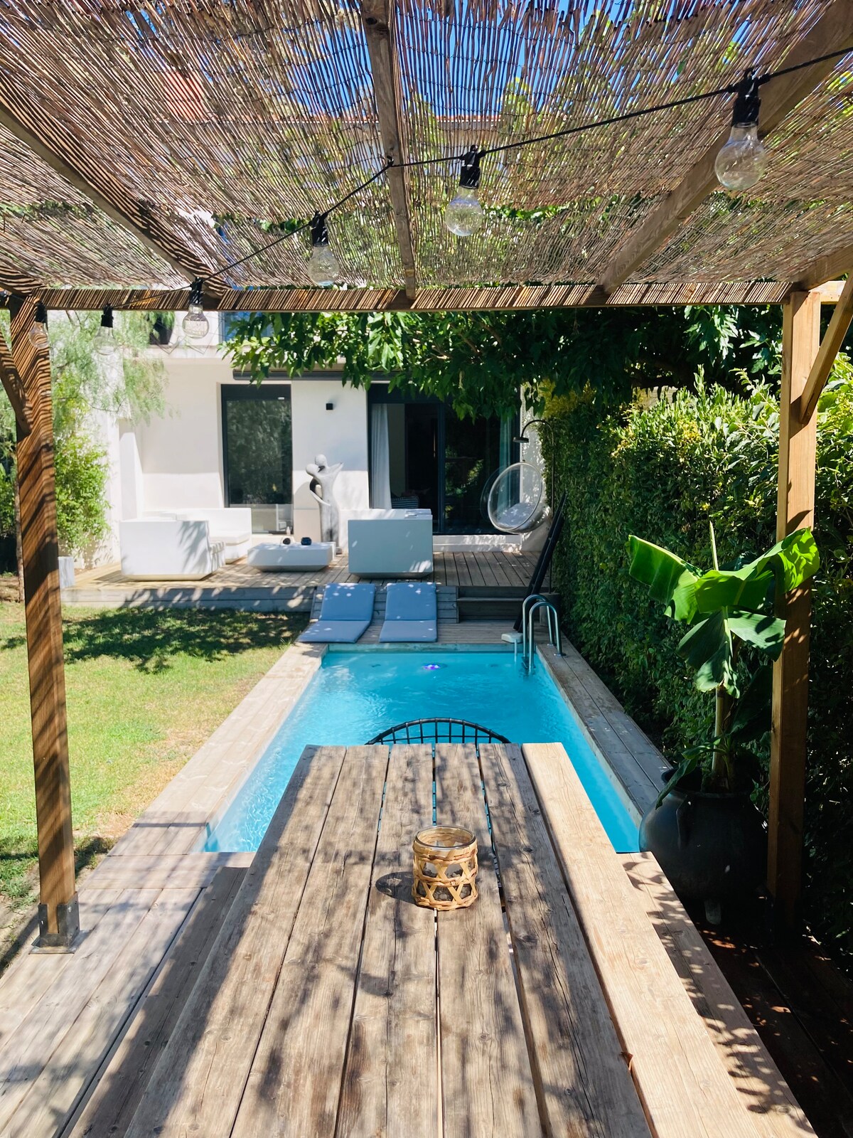 A wooden terrace with a table is visible, overlooking a small pool surrounded by greenery. Lounge chairs line the edge of the pool, and a nearby hanging chair is noted. The house features large windows, allowing natural light to enter the interior.
