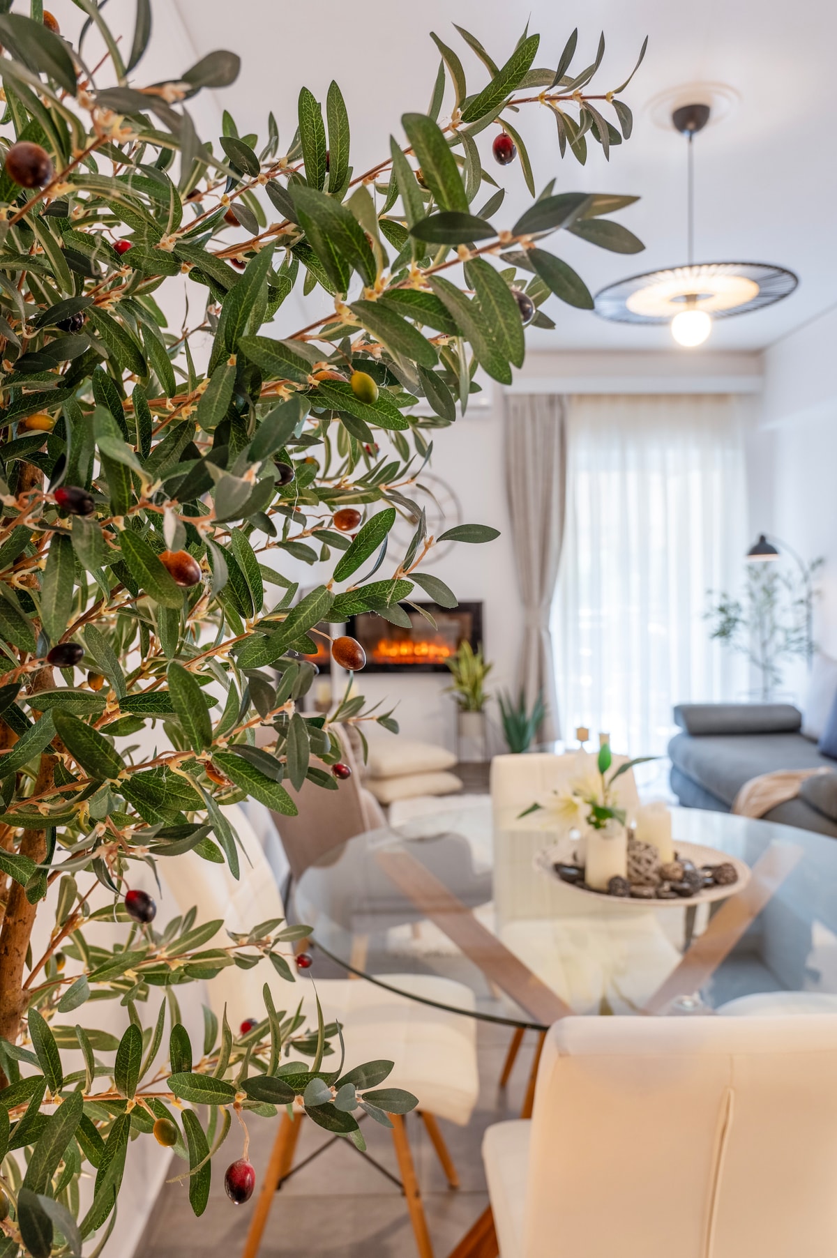 A dining area is depicted with a glass table surrounded by white chairs. An olive tree with dark green leaves and small berries adds a natural touch. A cozy living space is visible in the background, featuring a comfortable sofa and large windows with sheer curtains.